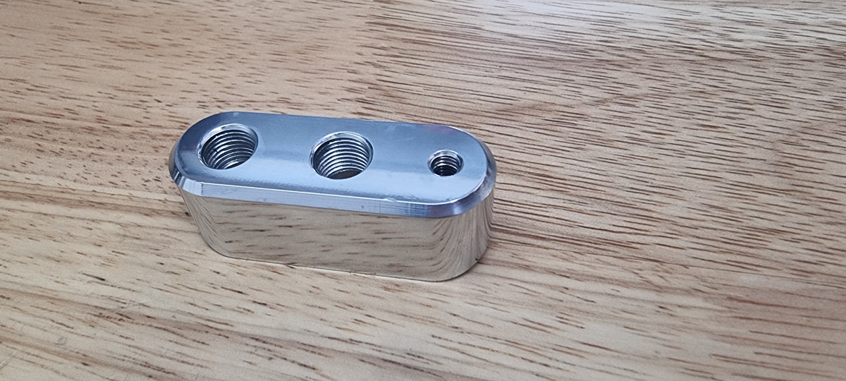 A silver billet weld-on boss made of 6061 aluminum with multiple ports for piping systems, placed on a wooden surface.