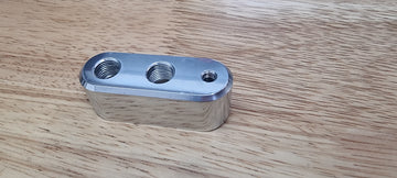 A silver billet weld-on boss made of 6061 aluminum with multiple ports for piping systems, placed on a wooden surface.