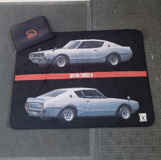 A black picnic blanket with a limited edition print of the Ken & Mary Skyline 2000GT-R car, displayed on a floor mat with a small carry case Ken & Mary Skyline 2000GT-R  Towel