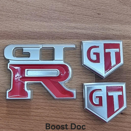 A set of custom OE-styled billet badges with anodised silver finish and red enamel accents, including 'GT' and 'GTR' emblems.