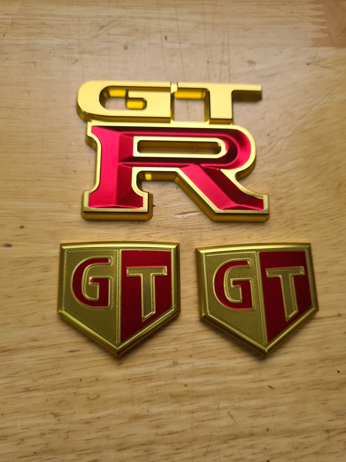 A set of GTR billet and titanium badges with red, gold, and black colors on a wooden surface.