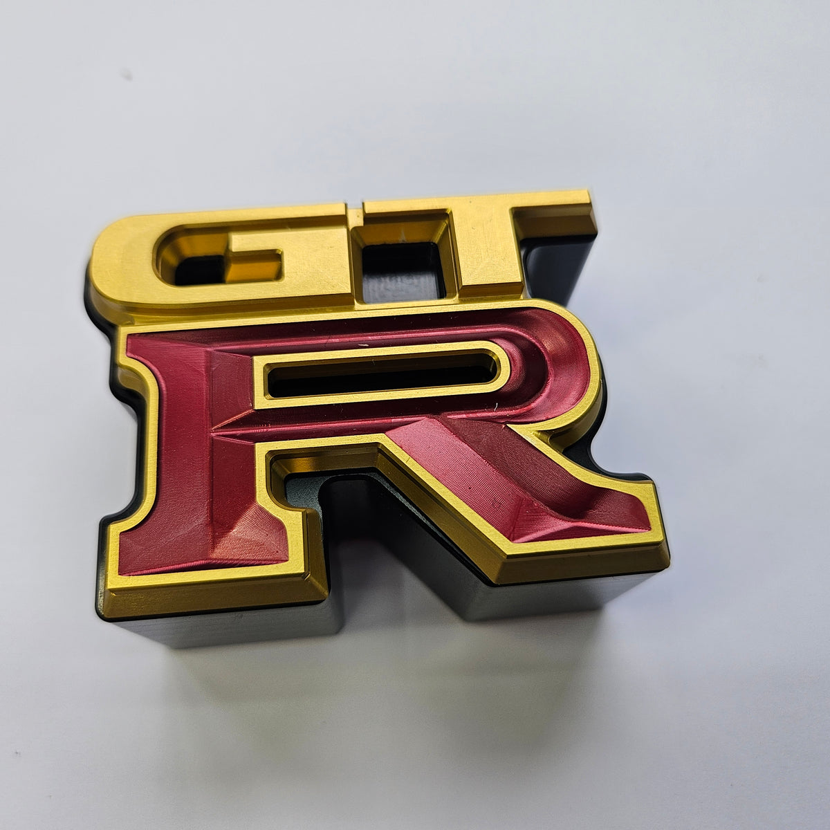 A 3D metallic letter 'R' emblem with gold edges and a deep red center, featuring a small 'GT' cutout at the top skyline replacement parts genuine oem nissan part